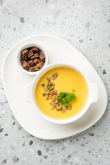 Creamy pumpkin soup in a white bowl, garnished with seeds and parsley, served with croutons on a white plate over a speckled gray background