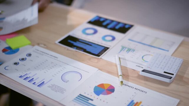A table with a bunch of graphs and a calculator. The graphs are of different sizes and colors, and the calculator is on top of them. Scene is focused and analytical, as the graphs