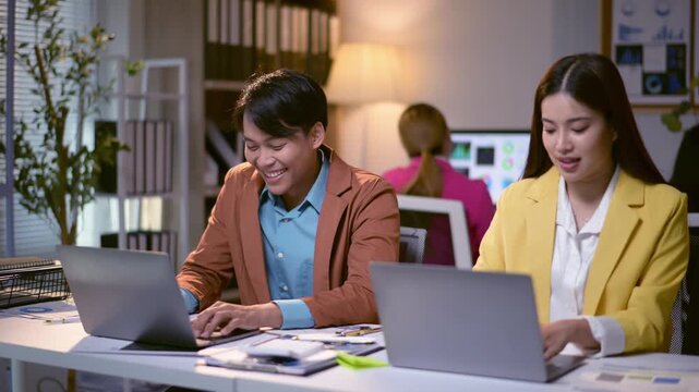 Two people are sitting at a desk with laptops open, one of them is wearing a yellow jacket