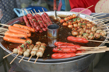 Horizontal photo of Thai night market street food, skewered sausages and meatballs served with a large pot of hot tamarind chili dipping sauce, authentic culinary scene.