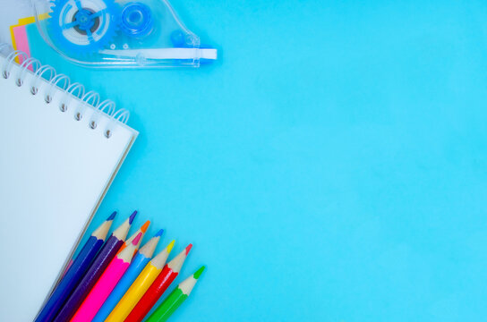 A bright blue background showcases a blank notebook and an array of colored pencils, along with a tape dispenser, inviting creativity and artistic expression. Copy space for your text.
