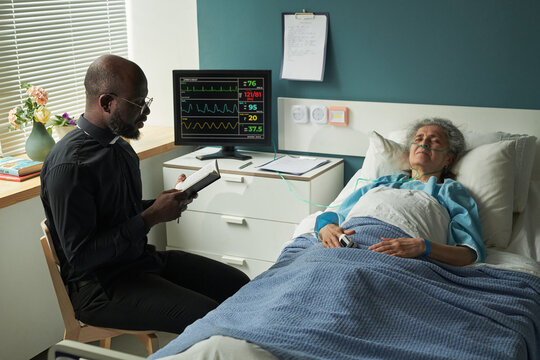 African American middle aged male priest sitting beside hospice bed reading prayers from Holy Bible to senior Caucasian woman resting with eyes closed, while providing end of life care
