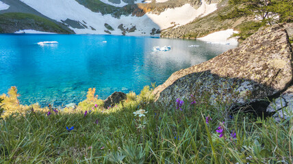 A serene mountain lake with clear blue water, snow-capped mountains, and rocky landscapes. Green...