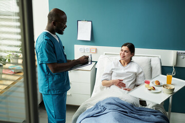 Caucasian young pregnant woman lying in maternity clinic bed talking to Blackmale nurse holding...