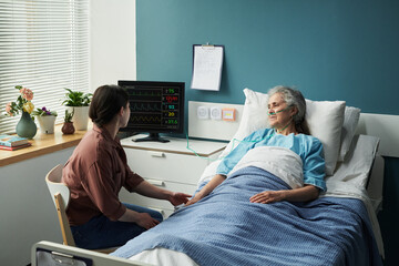Middle aged Caucasian woman sitting beside hospital bed holding hand of senior Caucasian woman lying under blanket with oxygen tube, medical monitor displaying vital signs nearby