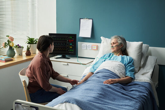 Middle aged Caucasian woman sitting beside hospital bed holding hand of senior Caucasian woman lying under blanket with oxygen tube, medical monitor displaying vital signs nearby