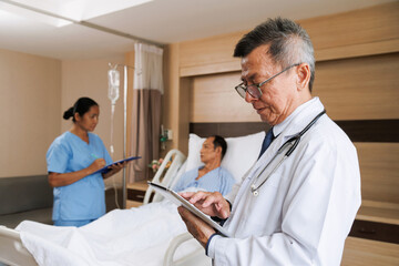 Obraz premium asian senior male professional doctor using tablet for digital health record and nurse writes notes beside patient. hospital room equipped medical instruments and IV drip. healthcare and treatment.