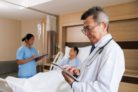 asian senior male professional doctor using tablet for digital health record and nurse writes notes beside patient. hospital room equipped medical instruments and IV drip. healthcare and treatment.