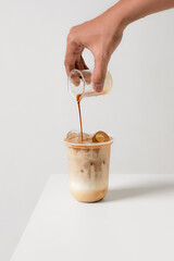 Hand pouring espresso into iced latte in a plastic cup on white background—minimal studio shot...