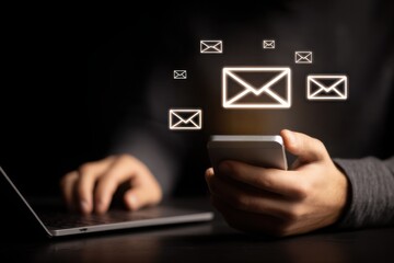 A person holding a phone and laptop  with email icons floating above
