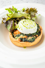 Vegetable tart with spinach filling, topped with poached egg and green sauce, served with fresh mixed greens on a white plate
