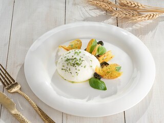 A ball of creamy burrata cheese rests on a white plate, surrounded by crispy bread slices, fresh...