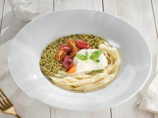 Delicate ribbons of pasta are arranged in a white bowl with creamy burrata, bright green basil...