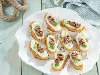 Golden slices of toasted baguette are arranged on a white plate and topped with creamy cheese,...