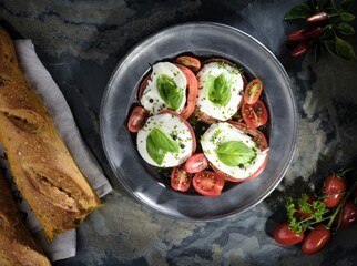 Caprese salad featuring sliced tomatoes, fresh Mozzarella, and basil sits on a rustic plate next to...
