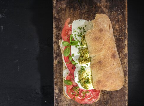 A vibrant Caprese sandwich features layers of fresh tomato, mozzarella, and basil. Pesto adds a flavor boost, all nestled in crusty bread atop a weathered wooden board