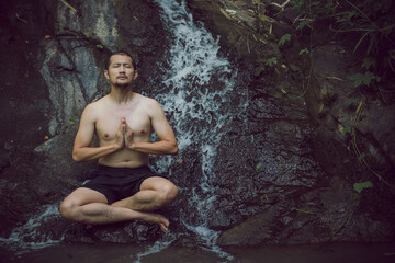 A Young Asian man in lotus yoga pose meditating on a peaceful natural waterfall. Beautiful natural sculpture of art and religion.
