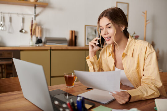 Bright and cheerful young woman engages in a phone call while reviewing important papers at home. - Powered by Adobe