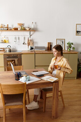 A young beautiful woman sips tea at a cozy table, focused on her work amidst a serene home setting.
