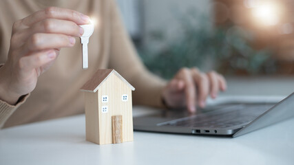 Hand holding a house key with a miniature wooden home and a laptop. Concept of online real estate,...