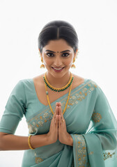 An elegant high-key portrait of an Indian woman in a teal-blue saree with a pleasant smile and hands clasped or folded hands