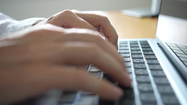 ノートパソコンでタイピングしている人。ナチュラルな色の木製のデスクで白いシャツを着た人がラップトップのキーボードを操作している。