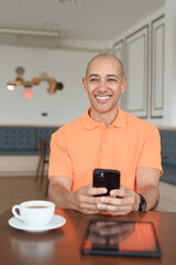 Bald Hispanic businessman having coffee break in cafe and using phone while thinking