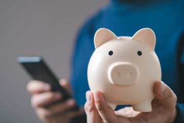 A person holds a piggy bank in one hand and a smartphone in the other, symbolizing saving and...