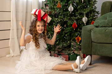 Cute funny little girl in an elegant fluffy white dress sits under Christmas tree, holding a gift...