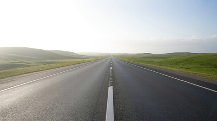 Fototapeta premium An empty asphalt road stretches into a bright hazy horizon with rolling green hills on either side under a pale sky