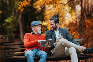 Senior father and his son sitting on bench by lake in nature, looking at tablet.