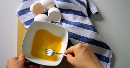 Closeup of hand whisking egg mixture in white bowl with fork, striped kitchen towel and egg shells...