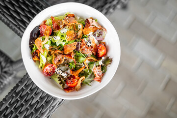 Fresh salad with mixed greens, cherry tomatoes, crispy bacon, sesame seeds, and creamy dressing in a white bowl on a wicker surface, top view
