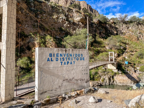 Willkommensschild mit der Aufschrift Bienvenidos al Distrito de Tapay vor einer H&auml;ngebr&uuml;cke