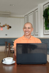 Bald Hispanic man smiling and using laptop computer in coffee shop restaurant while thinking