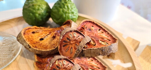 Dried and thinly sliced bael fruit is stacked on a wooden tray next to kaffir lime fruits. Sliced...