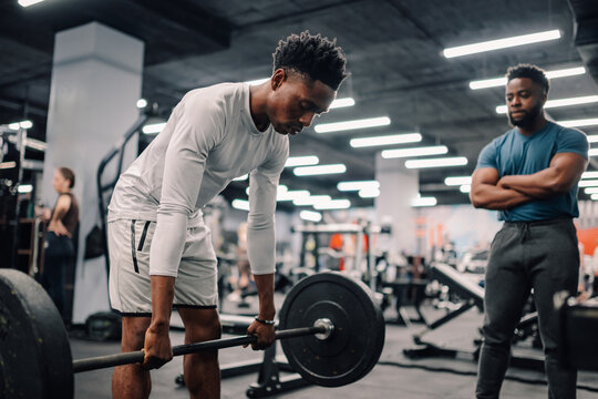 Determined young athlete lifting weights while personal trainer supervises in gym