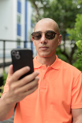 Bald Hispanic man face sitting outdoors at cafe restaurant wearing orange polo shirt using phone