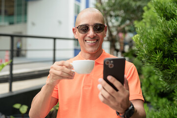 Bald Hispanic man face sitting outdoors at cafe restaurant wearing orange polo shirt using phone