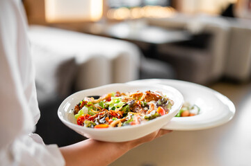 Close-up of a person holding a bowl of fresh salad with lettuce, cherry tomatoes, carrots, and grilled chicken in a cozy restaurant setting