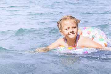 Young girl with blonde hair enjoying summer fun, floating on colorful inflatable ring in clear blue...