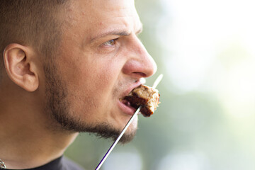 Man with short hair and beard, intensely biting into grilled meat on a skewer, showcasing a moment...