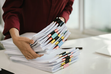 A cluttered office desk filled with paperwork, documents, files, and reports, symbolizing heavy...