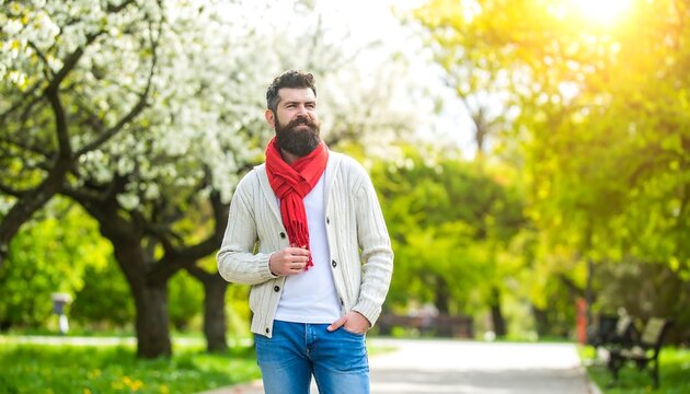 A man with a beard and red scarf strolls through a blossoming park on a sunny spring day.