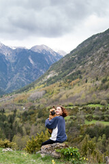 Naklejka premium Happy traveling woman with cute dog in highlands. Side view of content female hiker sitting on rock and tossing French Bulldog during adventure in Pyrenees mountain range