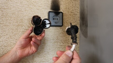 Male hands showing burnt wire with plug and tee against burnt socket on brown wallpapered wall, consequences of small fire due to electrical faults.