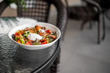 Fresh salad with poached egg, cherry tomatoes, and fried onions served in a white bowl on a glass table in an outdoor patio setting.