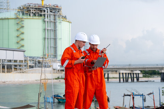Two workers in orange overalls consult digital tablet at industrial site near water under cloudy sky