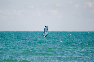 A windsurfer rides across vibrant blue-green waters beneath a clear sky with scattered clouds.