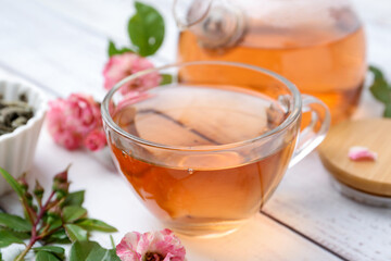 Tasty rose tea in glass cup, flowers and leaves on white wooden table, closeup